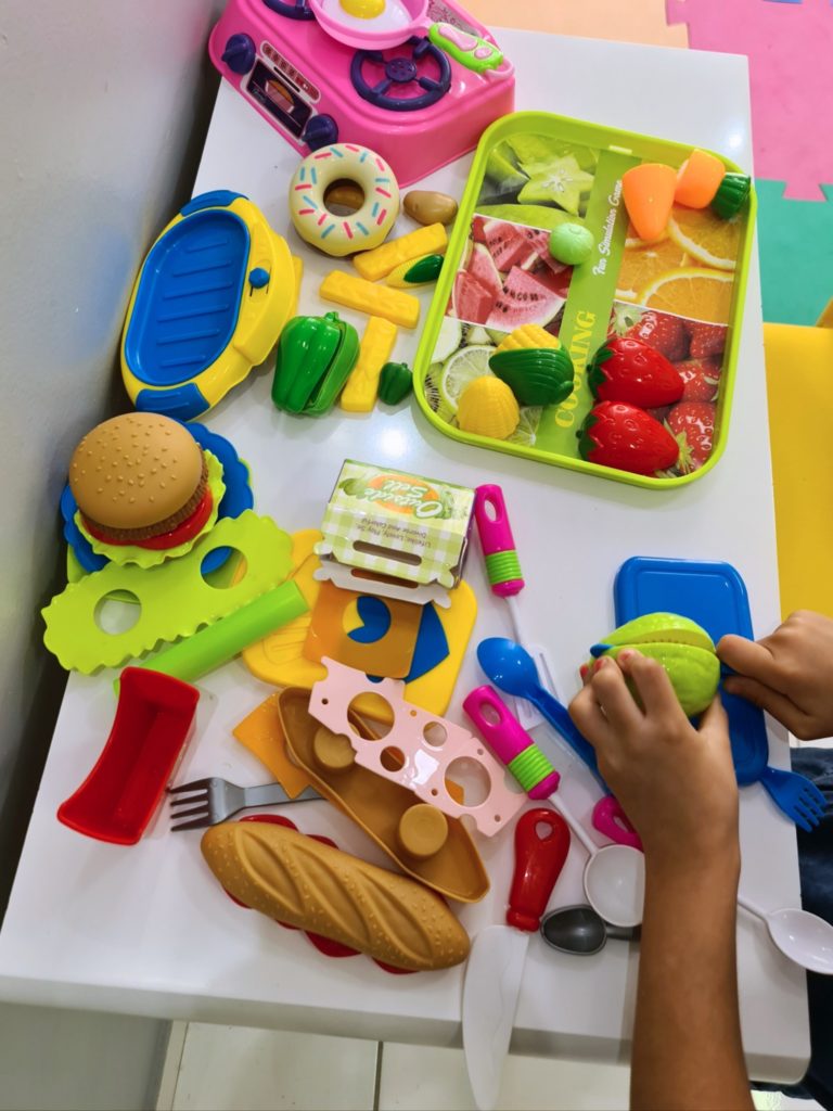 Ângela Berg: foco em mão de criança brincando com frutas e legumes de brinquedo